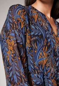 Tissu à motif floral bleu et orange sur un fond sombre. Design à encolure en V avec des manches fluides, mettant en valeur des motifs de feuilles vibrants.