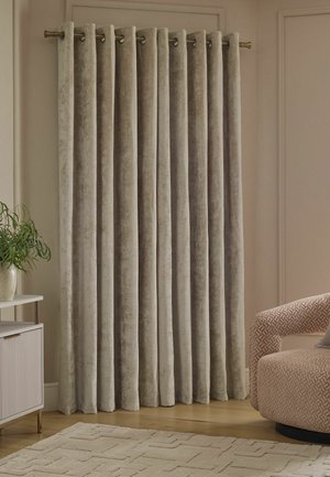 Tende di velluto beige appese a un’asta metallica in una stanza neutra con una poltrona a motivo, una piccola pianta e un tappeto testurizzato su pavimento in legno.