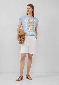 Jasnoniebieska koszulka graficzna z wzorem palmy, białe szorty z mankietem i brązowe klapki, trzymając brązową plecioną torbę.