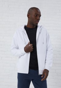 White zip-up hoodie with a drawstring hood, worn over a black shirt, featuring a smooth texture and ribbed cuffs, set against a white brick background.