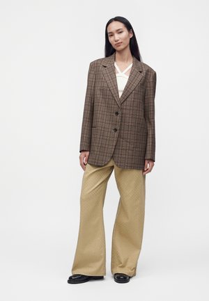 Femme aux longs cheveux foncés porte un blazer oversize marron à carreaux, un top blanc, un pantalon large beige à petits pois et des chaussures noires, debout sur fond blanc.