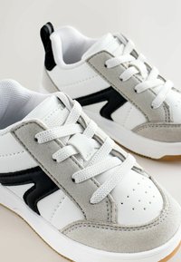 Baltos ir pilkos sportinės bateliai, turintys zomšos ir odos viršutinę dalį, juodus akcentus, apvalius raiščius ir tekstūruotą gumos pado.