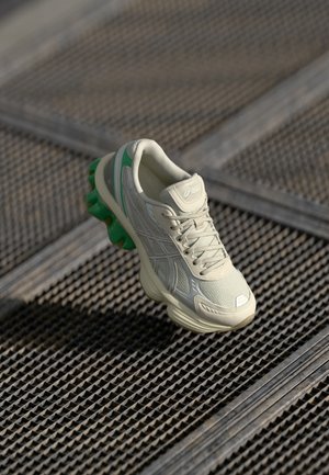 Jasnobeżowy but do biegania z zielonymi akcentami na podeszwie, spoczywający na teksturowanej metalowej powierzchni z wzorami siatki.