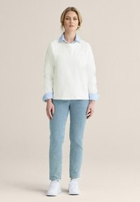 Vit långärmad top i lager över en ljusblå skjorta med krage, tillsammans med ljusblå jeans och vita sneakers, mot en neutral bakgrund.