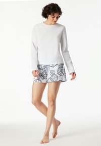 Vit långärmad topp med rund hals och subtil logotyp, matchad med en mönstrad skort med blå och grå design, ovanför knäet.