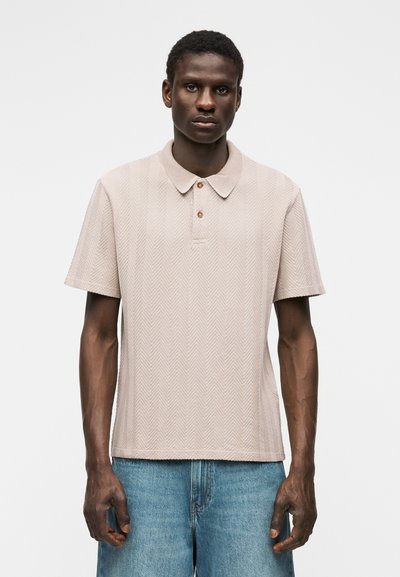 Άνδρας με μπεζ κοντομάνικο πόλο μπλουζάκι με σχέδιο σε σχήμα ψαροκόκαλου και ανοιχτό μπλε τζιν, στέκεται μπροστά σε απλό λευκό φόντο.