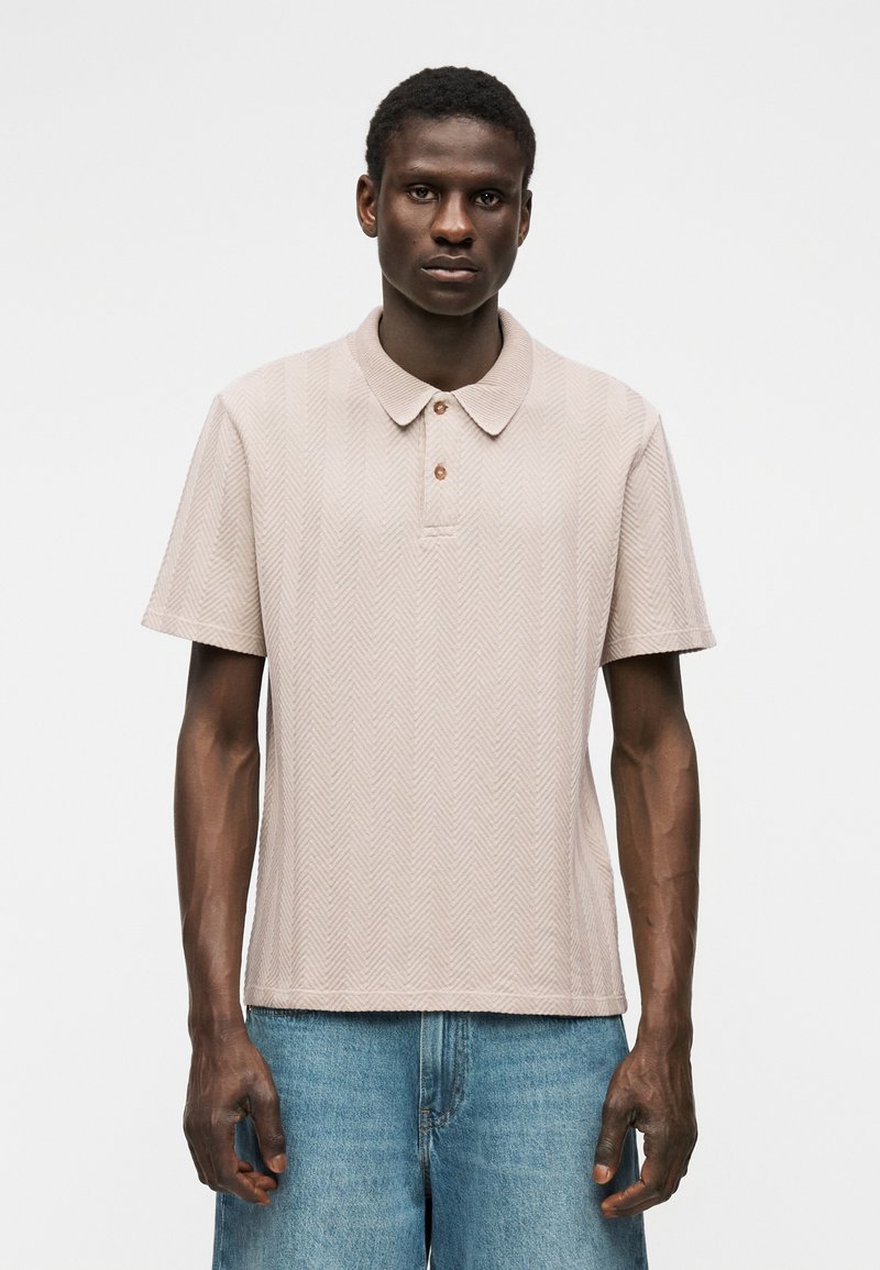 Homme portant un polo beige à manches courtes avec un motif chevron et un jean en denim bleu clair, debout devant un fond blanc uni.