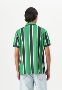 Male wearing green polo shirt with vertical navy and white stripes, paired with light blue denim jeans, standing facing white background.