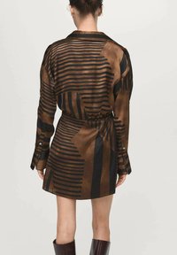Vrouw in een lange jurk met zwart en bruin gestreept abstract patroon en een taille die is geaccentueerd, gezien vanachter.
