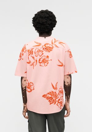 Henkilö, jolla on tummat kiharat hiukset ja joka pukeutunut vaaleanpunaiseen lyhythihaiseen paitaan, jossa on oranssinvärinen kukkakuosi, nähtynä takaa yksiväristä taustaa vasten.