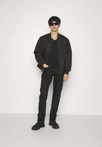 Uomo che indossa un giubbotto trapuntato nero, jeans neri, stivali neri e occhiali da sole, in piedi contro uno sfondo bianco uniforme.