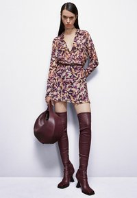 Abito floreale a maniche lunghe in viola, lavanda e crema con vita stretta, abbinato a stivali in pelle bordeaux alti fino alla coscia e una borsa coordinata.