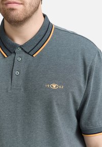 Polo gris en tissu texturé, avec une patte de boutonnage à deux boutons, des rayures de col contrastantes bleu marine et jaune, et un emblème avec "1962".