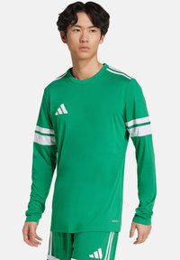 T-shirt de sport à manches longues vert, fabriqué en tissu lisse. Présente trois bandes blanches sur chaque manche et un logo Adidas sur la poitrine.