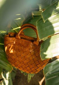 Woven tan leather handbag with rounded handles and a zipper closure. Surrounded by large green leaves, highlighting its texture and design.