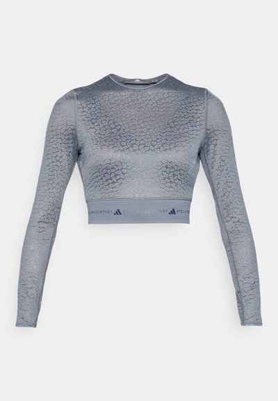 Κοντό μπλουζάκι με μακριά μανίκια σε ανοιχτό μπλε χρώμα με διακριτική υφή ζωηρού τύπου. Διαθέτει ζώνη μέσης με επωνυμία «Stella McCartney» και λογότυπο Adidas.