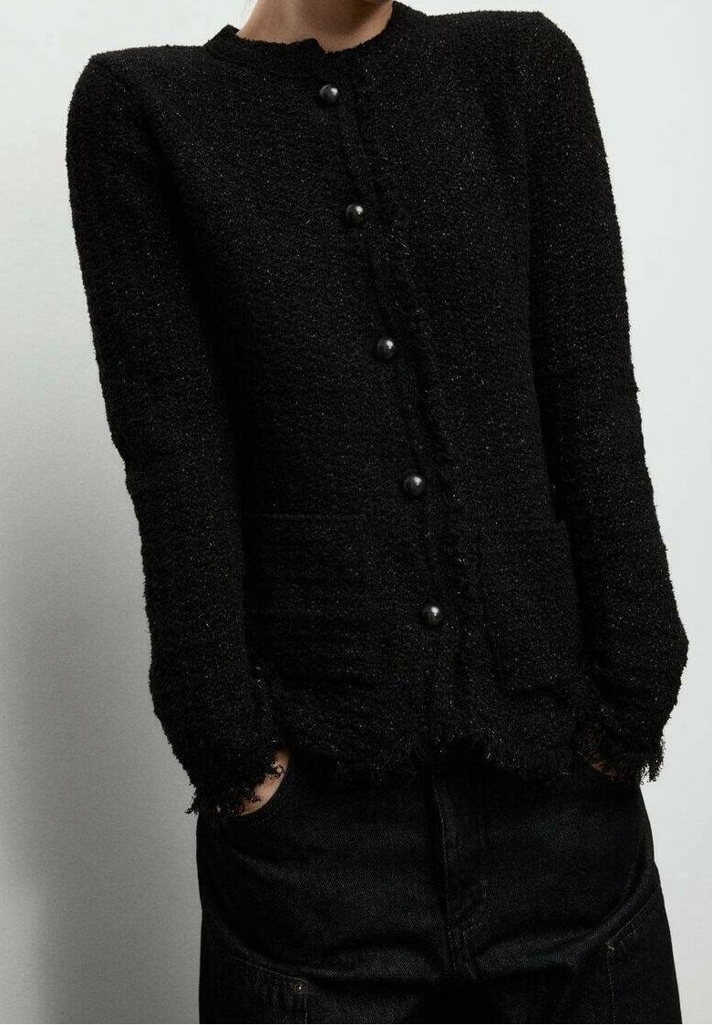 Personne portant un cardigan noir texturé boutonné avec manches longues et bords effilochés, associé à un pantalon noir, mains dans les poches.