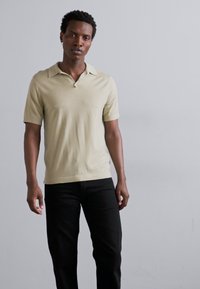 Ljust beige kortärmad polo skjorta med mjuk textur och en modest krage, matchad med mörka svarta slim-fit byxor.