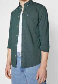 Man in een donkergroen overhemd met knopen over een wit T-shirt, met lichtblauwe jeans, een zilveren ring aan de linkerhand, en opgestroopte mouwen.