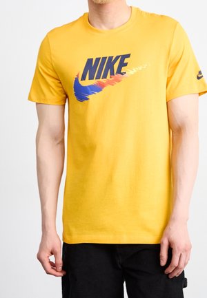 Mann in einem hellgelben Nike-T-Shirt mit einem verzerrten blauen und orangefarbenen Swoosh-Logo und schwarzer Hose, steht vor einem einfarbigen Hintergrund.