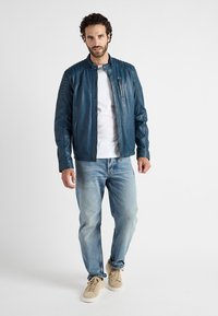 Heren blauwe leren jas met gewatteerde schouderaccenten en een hoge kraag. Rits borstzak en twee zijzakken. Gecombineerd met lichte jeans.