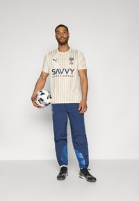 Bēšā svītrota sporta krekls ar tumši zilu logo, apvienots ar tumši zilām biksēm, kurās ir zilu akcentu. Tur rokās melnu un baltu futbola bumbu.