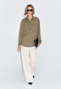 Olive green button-up shirt with pointed pockets, worn with white wide-leg trousers and a black handbag. Model poses against a light background.