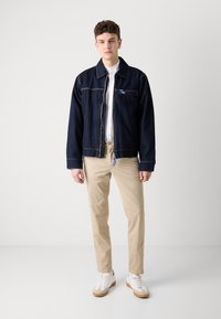 Tumša zilā džinsu jaka ar rāvējslēdzēju priekšā, krūšu kabatu un baltu šuvumu. Apvienota ar gaišām khaki biksēm un baltām kedām.