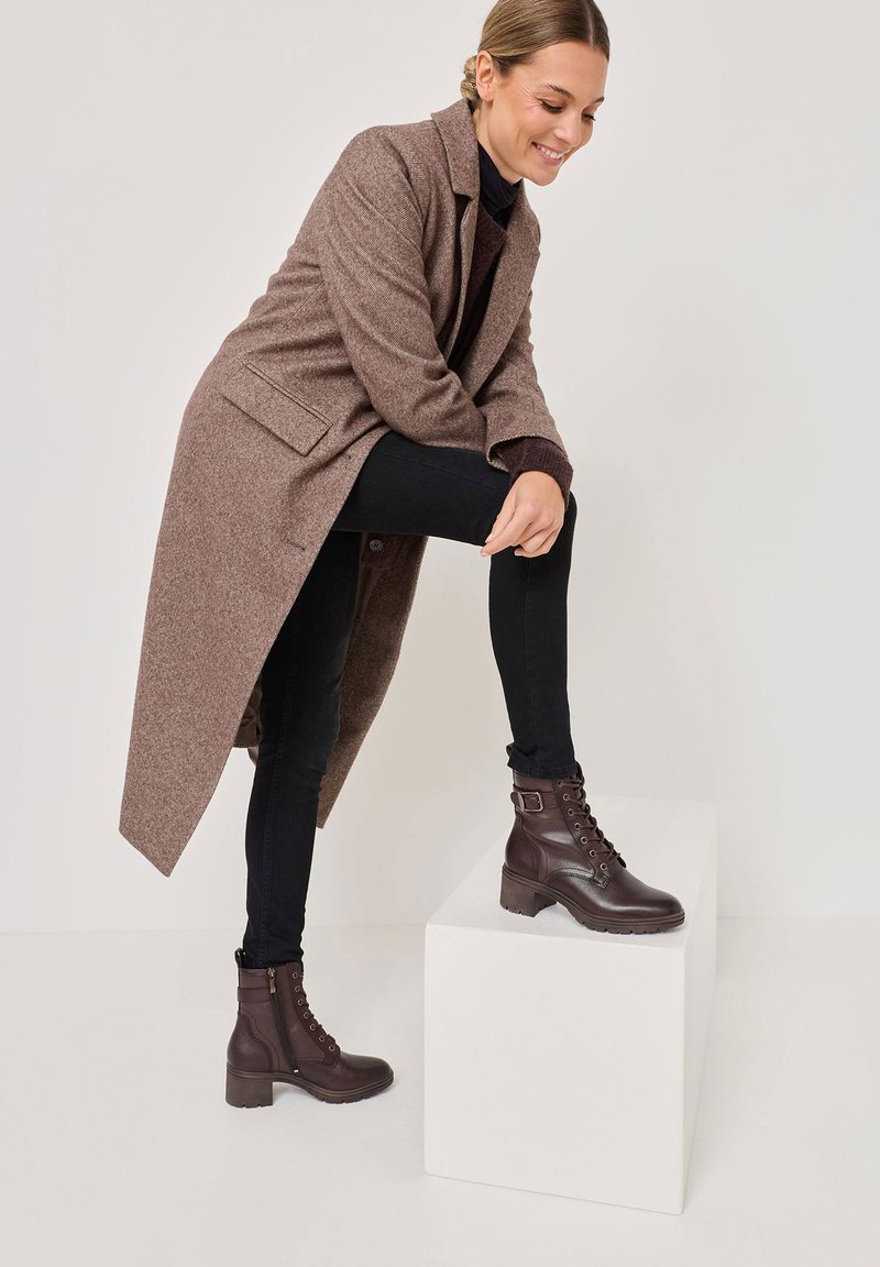 Hnědý vlněný kabát, černé skinny džíny a hnědé šněrovací boty s přezkou na bílém podstavci. Hladká textura, strukturovaný design a kontrastní barvy.