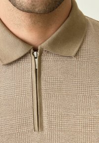 Polo beige con una cremallera corta, patrón texturizado y cuello plano. La tela tiene un sutil diseño a cuadros para añadir interés visual.