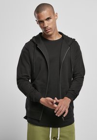 Sudadera negra con cremallera y cordones, bolsillos frontales y puños acanalados, llevada sobre una camiseta negra ajustada, combinada con pantalones verdes.