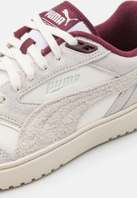 Zapatilla blanca y beige con materiales de ante y cuero, que presenta un panel lateral texturizado, acentos burdeos y un prominente logo de Puma.