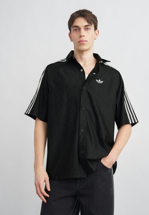 Νέος άνδρας που φοράει ένα μαύρο κοντομάνικο πουκάμισο Adidas με λευκές ρίγες στα μανίκια και λογότυπο στο στήθος, με το χέρι στην τσέπη.