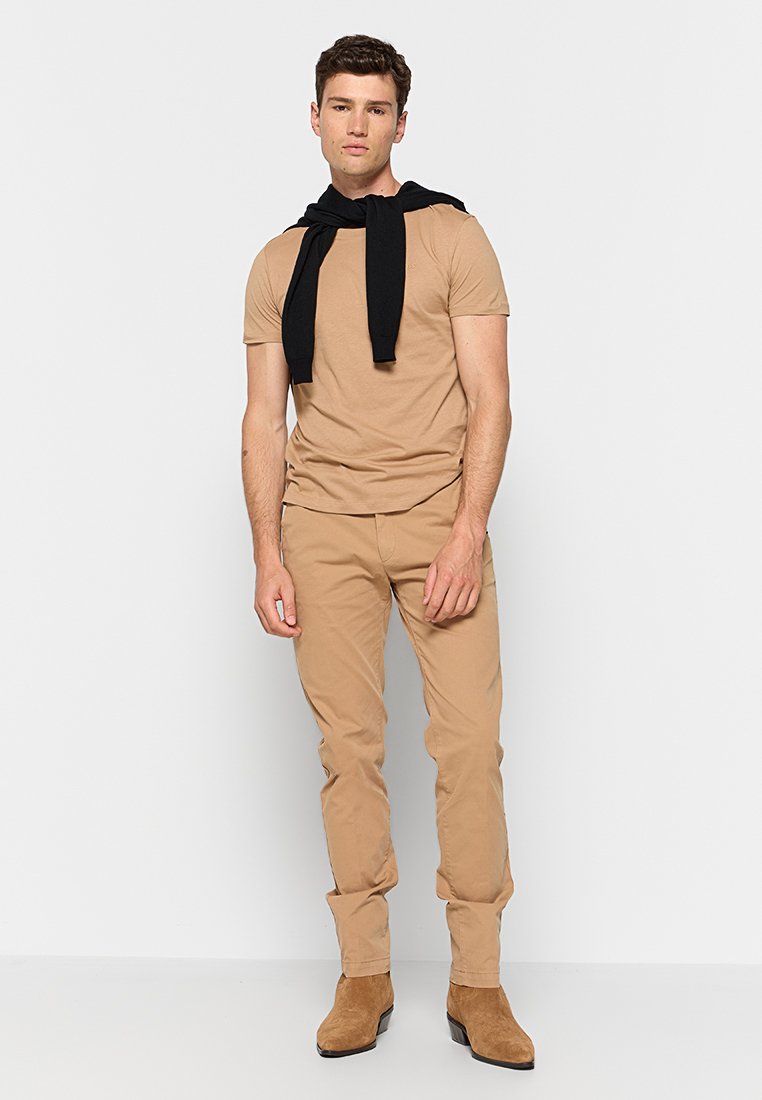 Junger Mann steht, trägt ein beiges kurzärmliges Hemd und Hose, schwarzer Pullover über den Schultern drapiert, beiges Wildlederstiefel auf weißem Hintergrund.