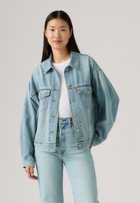 Veste en denim bleu clair avec une coupe décontractée, fermeture à boutons et poches poitrine. Portée par-dessus une chemise blanche et assortie avec un jean en denim.