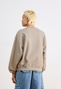 Hanorac supradimensionat de culoare taupe deschis, cu mâneci lungi, mansete cu nervuri și decolteu rotund. Are o textură netedă și un fit relaxat.