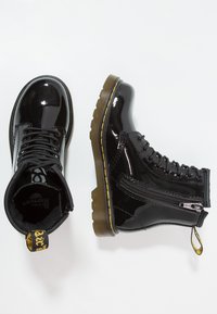 Botas de tobillo de charol negro con cordones en la parte delantera, cremallera lateral, costuras amarillas y suela de goma marrón resistente.