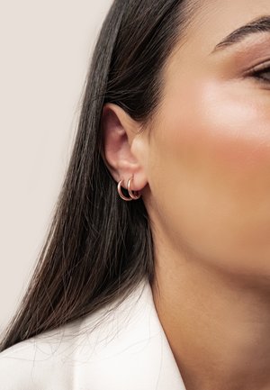 Nahaufnahme eines Frauenohrs mit zwei goldenen Creolen und dunklem, glattem Haar vor einem hellen, neutralen Hintergrund.