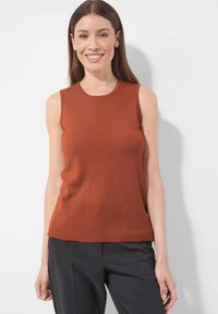 Frau in ärmellosem, rostfarbenem Oberteil und schwarzen Hosen, lächelt und steht vor einem einfarbigen, hellen Hintergrund.