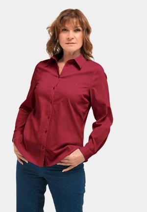 Rotes Hemd mit Knopfleiste, spitzen Kragen, langen Ärmeln und glatter Textur. Kombiniert mit dunkelblauen Jeans.
