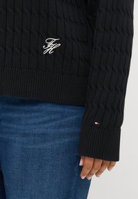 Čierny pletený sveter so vzorom káblov, s rebrovanými manžetami a vyšitým logom. V kombinácii s modrými džínsami, ktoré majú malý trojfarebný akcent na manžete.