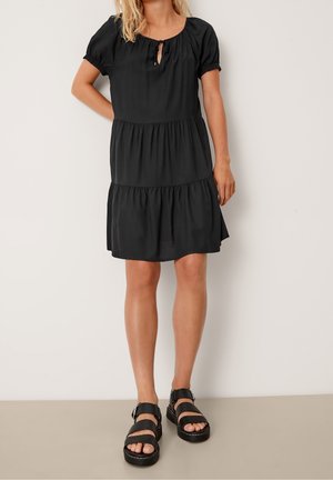 Femme portant une robe courte noire à volants avec des manches bouffantes et un nœud à l'avant, associée à des sandales plateformes noires, se tenant contre un mur clair.