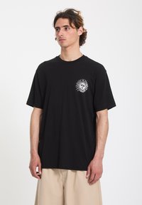 Giovane uomo che indossa una maglietta nera con una grafica di sole bianco e pantaloni beige, in piedi contro uno sfondo bianco uniforme.