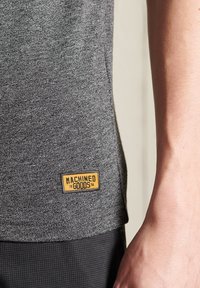 Primer plano de un dobladillo de camisa gris jaspeada con una etiqueta amarilla "Machined Goods" que lleva puesta una persona con un antebrazo visible.