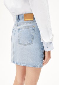 Jupe en denim bleu clair à coupe droite, avec poches arrière et un onglet en cuir marron au niveau de la taille. Les finitions comprennent une surpiqûre.