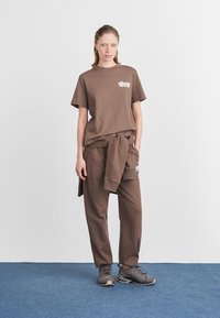 Brun bomulls t-skjorte med korte ermer og hvit logo; matchende brune joggebukser; modellen har på seg grå treningssko; ensfarget bakgrunn.
