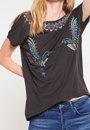 Camiseta negra con colibríes bordados en colores vivos y patrones geométricos cerca del escote, combinada con pantalones vaqueros azules.