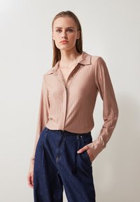 Light pink, long-sleeve blouse featuring vertical ribbing, button-down front, and a classic collar. Paired with dark blue high-waisted trousers.