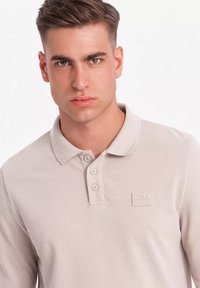 Polo shirt color beige chiaro con un motivo testurizzato, tre bottoni e un logo in rilievo sul lato sinistro del petto. Il tessuto sembra morbido e traspirante.