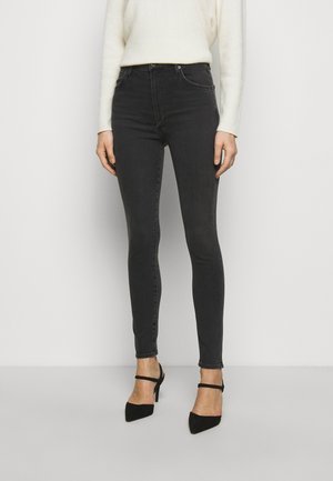 Kobieta ubrana w czarne, obcisłe jeansy z wysokim stanem, biały sweter oraz czarne szpilki z zakrzywionym czubkiem i paskami wokół kostek.